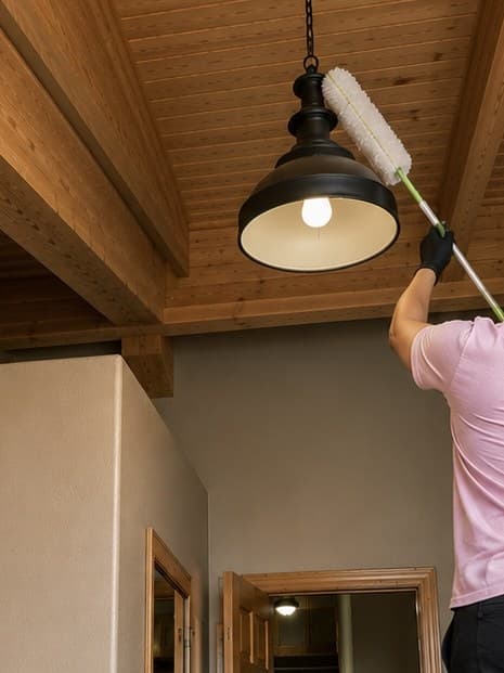 A ShinySpaces cleaner using an extended duster to clean a high pendant light fixture as part of a custom cleaning solution